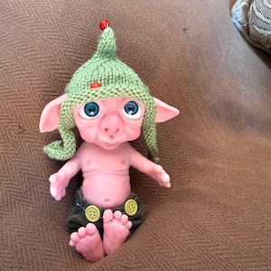 May include: A small, pale pink creature figurine with large, bright blue eyes and pointed ears. It wears a green knitted pointed hat with two small red beads and dark olive-colored pants featuring two visible yellow buttons at the waistband.