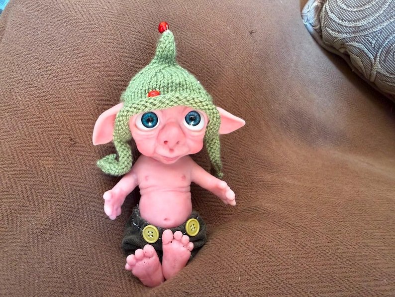 May include: A small, pale pink creature figurine with large, bright blue eyes and pointed ears. It wears a green knitted pointed hat with two small red beads and dark olive-colored pants featuring two visible yellow buttons at the waistband.