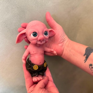 May include: A small, pink, fantasy-style creature figurine with large, bright blue eyes, prominent ears, and a smooth, rounded head. The figure has a small torso and is wearing dark brown fabric shorts that are fastened with two yellow buttons.