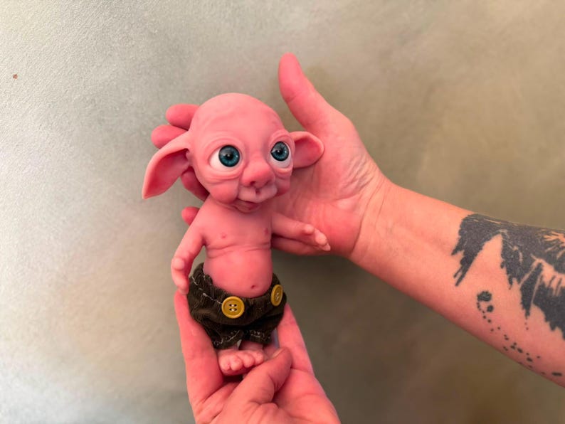 May include: A small, pink, fantasy-style creature figurine with large, bright blue eyes, prominent ears, and a smooth, rounded head. The figure has a small torso and is wearing dark brown fabric shorts that are fastened with two yellow buttons.