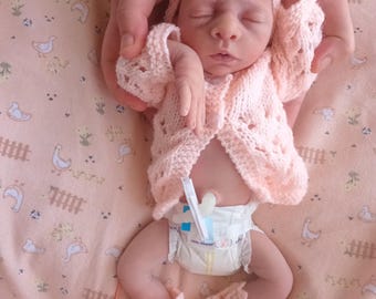 May include: A newborn baby doll wearing a light pink knitted outfit, including a hat with a bow, a cardigan, and booties with ribbons. The baby is lying on a pink patterned blanket.
