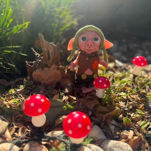 May include: A small fantasy creature doll with large blue eyes, wearing a green knit cap, a brown vest with a small orange heart patch, and dark pants. The doll stands near a tree stump surrounded by several red and white spotted mushroom ornaments.
