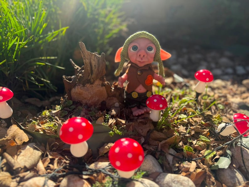 May include: A small fantasy creature doll with large blue eyes, wearing a green knit cap, a brown vest with a small orange heart patch, and dark pants. The doll stands near a tree stump surrounded by several red and white spotted mushroom ornaments.