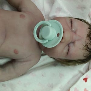 May include: A realistic baby doll with closed eyes and a pacifier. The doll has a fair skin tone and detailed facial features. The pacifier is light green. The baby is lying on a white and pink patterned blanket.