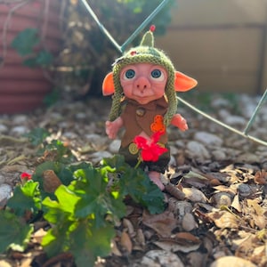 May include: A small, handcrafted doll with large blue eyes, pointed ears, and a pinkish complexion. It wears a textured green knit hat with a small red accent, a brown vest with a bright orange patch, and dark green trousers with gold-colored buttons.