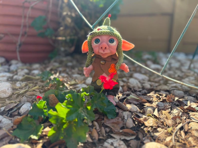 May include: A small, handcrafted doll with large blue eyes, pointed ears, and a pinkish complexion. It wears a textured green knit hat with a small red accent, a brown vest with a bright orange patch, and dark green trousers with gold-colored buttons.