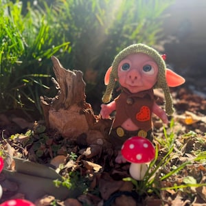 May include: A small fantasy creature figurine with large ears and eyes, wearing a green knit hat, a brown vest with an orange heart patch, and dark shorts with yellow buttons, standing near a piece of driftwood and several red and white mushrooms.