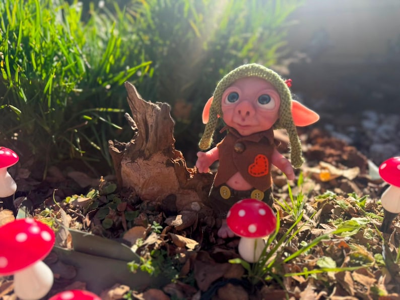 May include: A small fantasy creature figurine with large ears and eyes, wearing a green knit hat, a brown vest with an orange heart patch, and dark shorts with yellow buttons, standing near a piece of driftwood and several red and white mushrooms.
