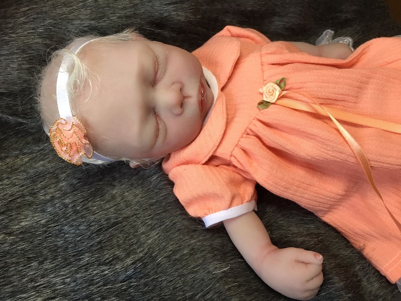 K&ouml;nnte beinhalten: Eine schlafende Baby-Puppe, die ein pfirsichfarbenes Kleid mit wei&szlig;em Kragen und wei&szlig;en &Auml;rmelb&uuml;ndchen tr&auml;gt. Die Puppe hat blondes Haar und tr&auml;gt ein wei&szlig;es Stirnband mit einer pfirsichfarbenen Blume.