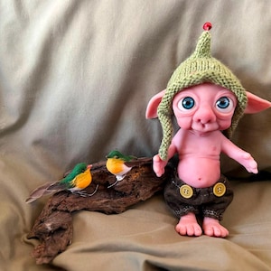 May include: A pink-skinned fantasy creature figurine with large blue eyes, wearing a green knitted hat with a red bead and brown shorts with two yellow buttons. Beside the creature, two small artificial birds rest on a piece of driftwood.