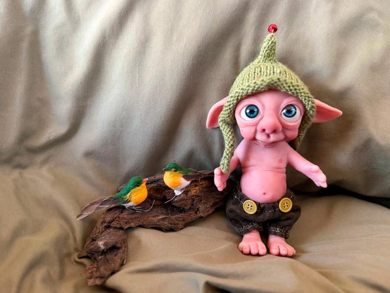 May include: A pink-skinned fantasy creature figurine with large blue eyes, wearing a green knitted hat with a red bead and brown shorts with two yellow buttons. Beside the creature, two small artificial birds rest on a piece of driftwood.
