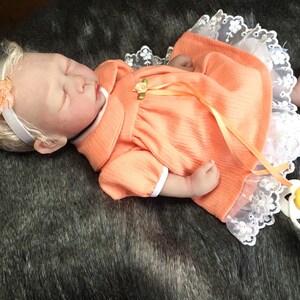 K&ouml;nnte beinhalten: Eine schlafende Reborn-Puppe, die ein pfirsichfarbenes Kleid mit wei&szlig;em Spitzenbesatz und einem gelben Band tr&auml;gt. Die Puppe hat blonde Haare und tr&auml;gt ein Stirnband mit einer pfirsichfarbenen Blume.