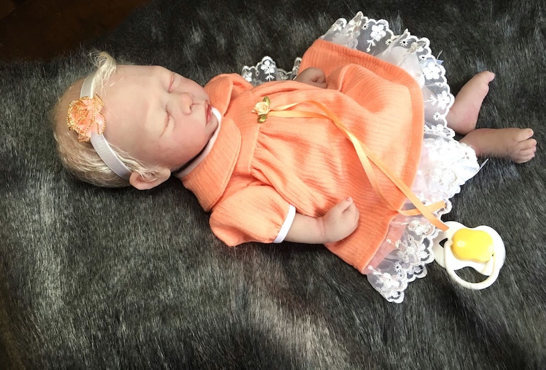 K&ouml;nnte beinhalten: Eine schlafende Reborn-Puppe, die ein pfirsichfarbenes Kleid mit wei&szlig;em Spitzenbesatz und einem gelben Band tr&auml;gt. Die Puppe hat blonde Haare und tr&auml;gt ein Stirnband mit einer pfirsichfarbenen Blume.