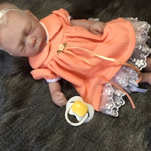 K&ouml;nnte beinhalten: Eine schlafende Reborn-Puppe, die ein pfirsichfarbenes Kleid mit wei&szlig;em Spitzenbesatz und einen gelben Schnuller tr&auml;gt. Die Puppe hat blonde Haare und tr&auml;gt ein Stirnband mit einer pfirsichfarbenen Blume.