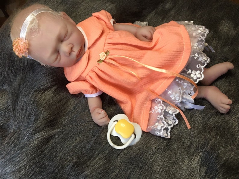 K&ouml;nnte beinhalten: Eine schlafende Reborn-Puppe, die ein pfirsichfarbenes Kleid mit wei&szlig;em Spitzenbesatz und einen gelben Schnuller tr&auml;gt. Die Puppe hat blonde Haare und tr&auml;gt ein Stirnband mit einer pfirsichfarbenen Blume.