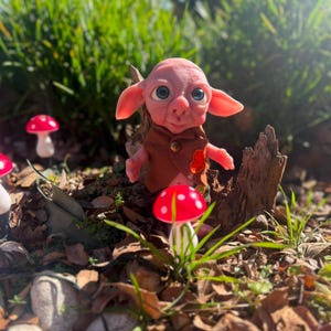 May include: A small, pink-skinned creature with large blue eyes and pointed ears, wearing a brown vest with a single button. The figure is surrounded by several small, artificial mushrooms with bright red caps and white stems in a garden setting.