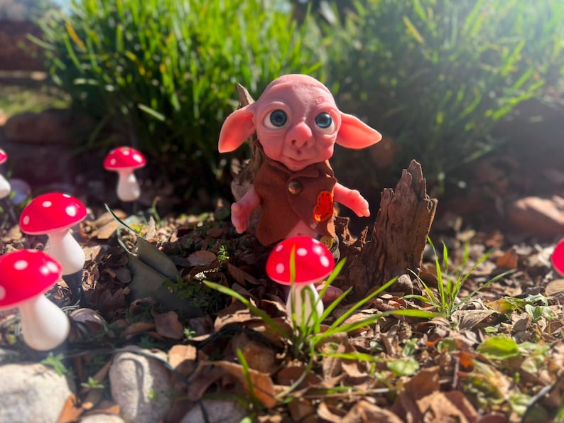 May include: A small, pink-skinned creature with large blue eyes and pointed ears, wearing a brown vest with a single button. The figure is surrounded by several small, artificial mushrooms with bright red caps and white stems in a garden setting.