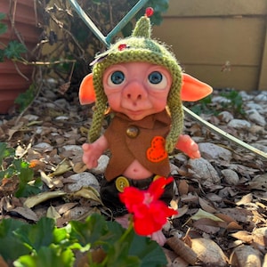 May include: A small fantasy creature doll with pink skin, large blue eyes, and pointed ears. It wears a green knitted hat with a small red bead, a brown felt vest with a brass button and orange heart patch, and dark brown pants with yellow buttons.