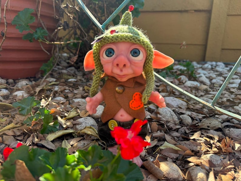 May include: A small fantasy creature doll with pink skin, large blue eyes, and pointed ears. It wears a green knitted hat with a small red bead, a brown felt vest with a brass button and orange heart patch, and dark brown pants with yellow buttons.