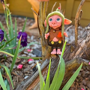 May include: A small fantasy creature figurine with pink skin, large blue eyes, and pointed ears. It wears a green knit cap with a red tip, a brown vest with an orange heart patch, and dark brown shorts featuring two yellow buttons on the front.