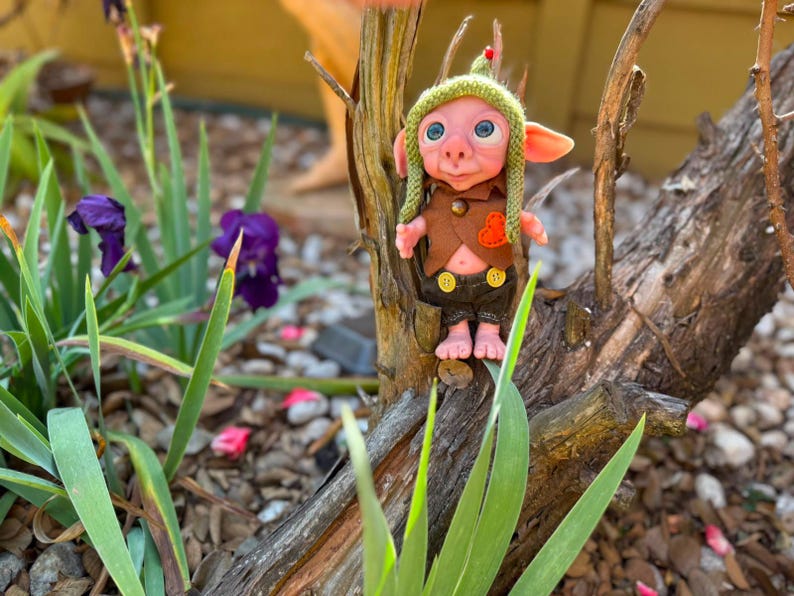 May include: A small fantasy creature figurine with pink skin, large blue eyes, and pointed ears. It wears a green knit cap with a red tip, a brown vest with an orange heart patch, and dark brown shorts featuring two yellow buttons on the front.