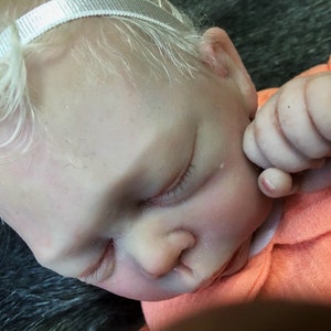 K&ouml;nnte beinhalten: Nahaufnahme einer schlafenden Reborn-Puppe mit blonden Haaren, die ein wei&szlig;es Stirnband und ein orangefarbenes Hemd tr&auml;gt. Die Augen der Puppe sind geschlossen und der Mund leicht ge&ouml;ffnet.
