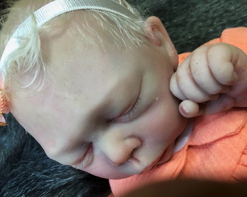 K&ouml;nnte beinhalten: Nahaufnahme einer schlafenden Reborn-Puppe mit blonden Haaren, die ein wei&szlig;es Stirnband und ein orangefarbenes Hemd tr&auml;gt. Die Augen der Puppe sind geschlossen und der Mund leicht ge&ouml;ffnet.