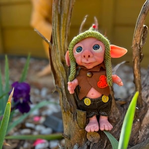 May include: A small fantasy creature doll with pink skin, large blue eyes, and pointed ears. It wears a green knit cap, a brown vest with a small orange heart patch, and dark brown shorts featuring two yellow buttons on the front waistband.