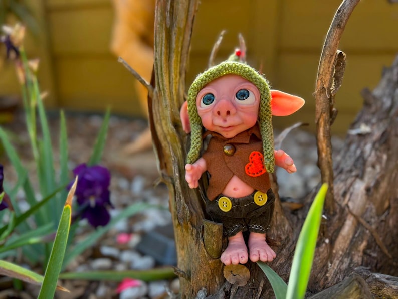 May include: A small fantasy creature doll with pink skin, large blue eyes, and pointed ears. It wears a green knit cap, a brown vest with a small orange heart patch, and dark brown shorts featuring two yellow buttons on the front waistband.