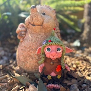 May include: A small, pink-skinned fantasy creature doll with large blue eyes, wearing a green knitted hat with a small red bead, a brown vest with a red heart patch, and dark pants with two yellow buttons, standing in front of a stone hedgehog.