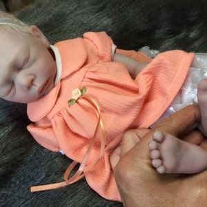 K&ouml;nnte beinhalten: Eine schlafende Reborn-Puppe, die ein pfirsichfarbenes Kleid mit wei&szlig;er Spitzenborte und einem rosa Band tr&auml;gt. Die Puppe hat ein wei&szlig;es Stirnband mit einer rosa Schleife.