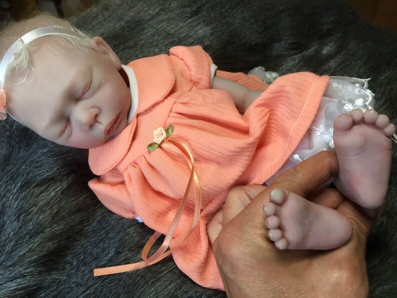 K&ouml;nnte beinhalten: Eine schlafende Reborn-Puppe, die ein pfirsichfarbenes Kleid mit wei&szlig;er Spitzenborte und einem rosa Band tr&auml;gt. Die Puppe hat ein wei&szlig;es Stirnband mit einer rosa Schleife.
