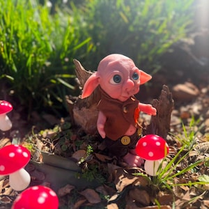 May include: A small, pink-skinned fantasy creature figurine with large blue eyes and pointed ears. The figure wears a brown vest with a gold button and dark brown shorts. It is positioned near several red and white spotted mushroom decorations.