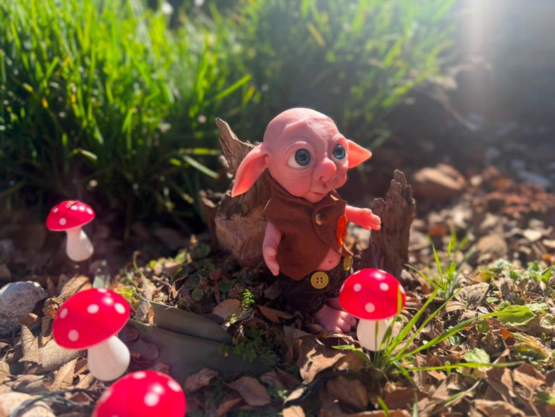 May include: A small, pink-skinned fantasy creature figurine with large blue eyes and pointed ears. The figure wears a brown vest with a gold button and dark brown shorts. It is positioned near several red and white spotted mushroom decorations.