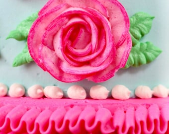 Puede incluir: Un pastel glaseado rosa y blanco con una rosa rosa y hojas verdes en la parte superior. El pastel está decorado con un borde de volantes rosa.