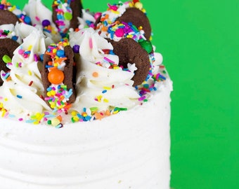 May include: A white frosted cake decorated with colorful sprinkles and chocolate sandwich cookies. The cake is on a pink cake stand.