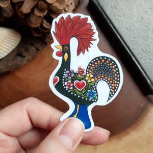May include: A colourful rooster sticker with a black body, red comb and wattle, and a tail with a pattern of colourful dots. The rooster is surrounded by a white border.