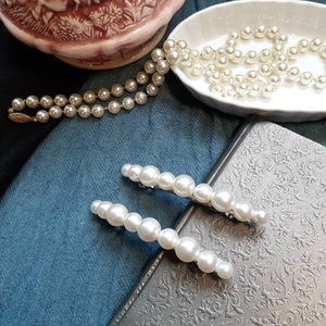 Two large pearl barrettes leaning on a charcoal book. In background you can see blue and black fabric and a porcelain bowl with a pearl necklace pouring out. On the top left a fancy pink pitcher.