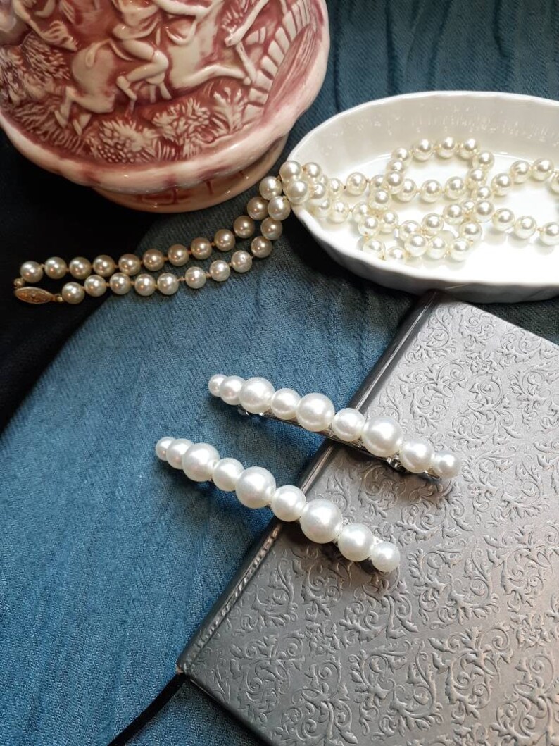 Two large pearl barrettes leaning on a charcoal book. In background you can see blue and black fabric and a porcelain bowl with a pearl necklace pouring out. On the top left a fancy pink pitcher.