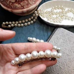One large pearl barrettes leaning on a charcoal book. The other in hand to see up close. In background you can see blue and black fabric and a porcelain bowl with a pearl necklace pouring out. On the top left a fancy pink pitcher.