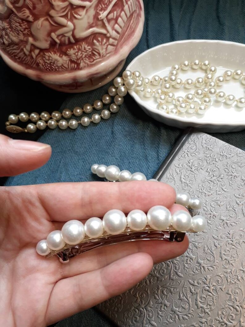 One large pearl barrettes leaning on a charcoal book. The other in hand to see up close. In background you can see blue and black fabric and a porcelain bowl with a pearl necklace pouring out. On the top left a fancy pink pitcher.