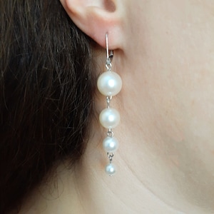 Peut inclure: Une paire de boucles d'oreilles pendantes en argent avec quatre perles blanches. Les perles sont rondes et de tailles différentes, la plus grosse étant en haut.