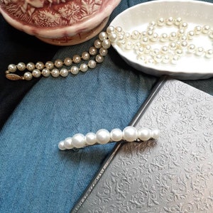 A large pearl barrette leaning on a charcoal book. In background you can see blue and black fabric and a porcelain bowl with a pearl necklace pouring out. On the top left a fancy pink pitcher.