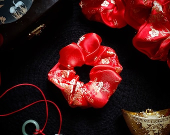 May include: Red satin hair scrunchies with gold floral patterns. The scrunchies are on a black surface with a small jade pendant and a gold decorative box.
