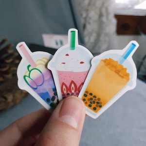 Könnte beinhalten: Drei bunte Bubble Tea-Aufkleber mit verschiedenen Geschmacksrichtungen. Die Aufkleber zeigen einen rosa Bubble Tea mit Erdbeeren, einen blauen Bubble Tea mit Mango und einen gelben Bubble Tea mit Tapiokaperlen.