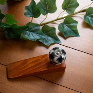 May include: A wooden door stop with a decorative knob. The knob is white with a black starburst pattern and a silver centre. The door stop is brown and sits on a wooden surface with green leaves in the background.