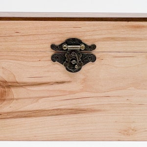 May include: A wooden box with a light-colored finish and a dark brown top. The box features a decorative metal clasp in the center. The wood grain is visible, adding to the natural aesthetic. This box is suitable for storage.