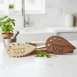 May include: Two wooden herb strippers, one light and one dark, with text labels for various herbs. The light stripper has "Oregano", "Sage", "Mint", "Parsley", "Dill", and "Thyme" labels. The dark stripper has similar labels. Both have a leaf design.