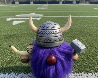 May include: A whimsical purple gnome figurine with a Viking helmet, holding a hammer and a horn. The gnome has a round red nose and a fluffy beard. The background is a green football field.