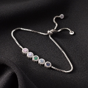 Könnte beinhalten: Ein silbernes Kettenarmband mit einem Schiebeverschluss und einer Reihe von sieben kleinen runden Steinen. Die Steine sind in Silber gefasst und haben verschiedene Farben, darunter Rosa, Lila, Grün, Blau und Klar.